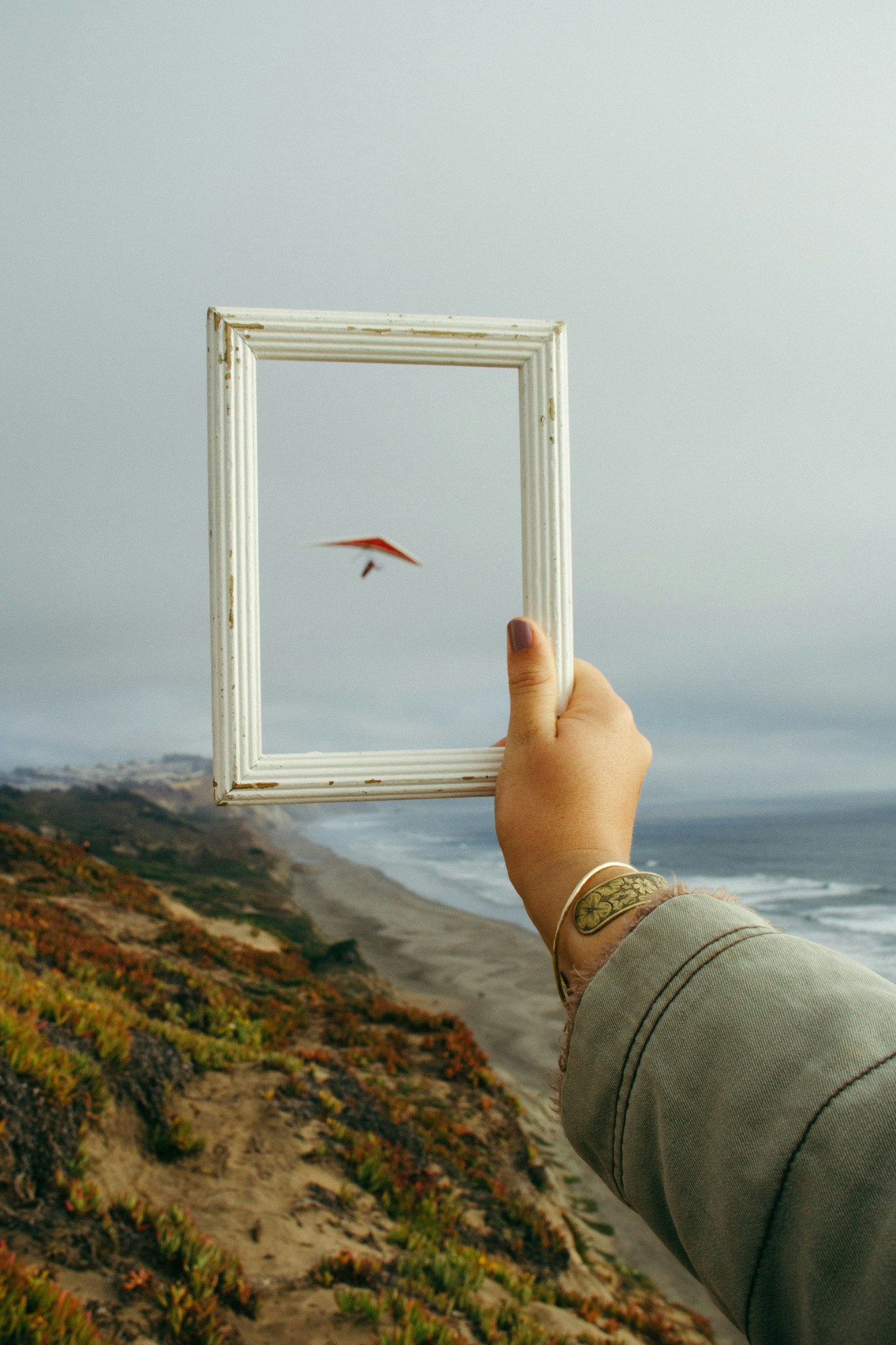 A hand holding up a white picture frame against a coastal cliff and ocean, with a hang glider flying through it